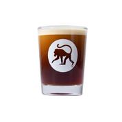 Frothy Monkey logo shot glass filled with espresso, white monkey emblem on clear thick-walled glass.