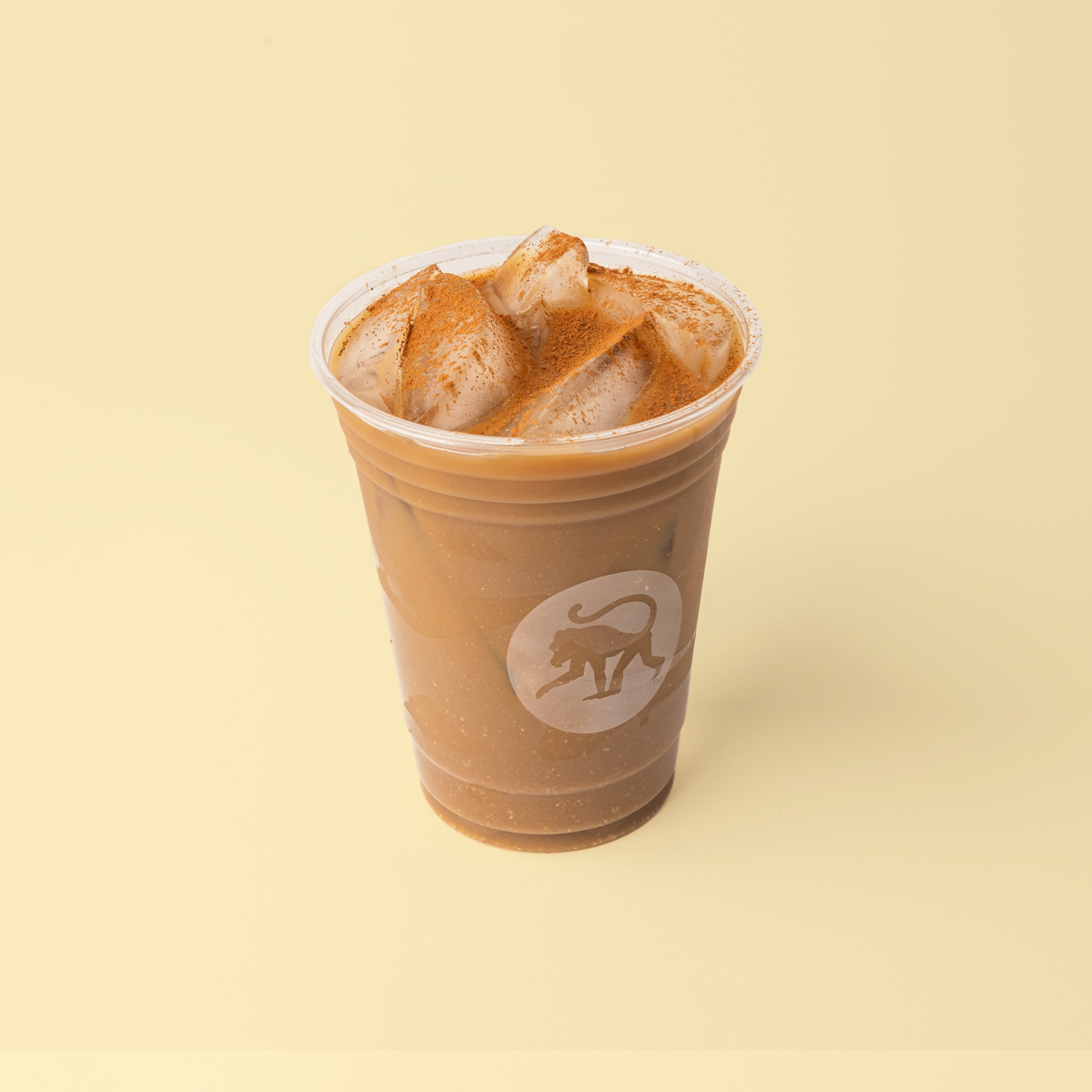 An iced latte in a clear Frothy Monkey cup topped with cinnamon-dusted ice, set against a pale yellow background.