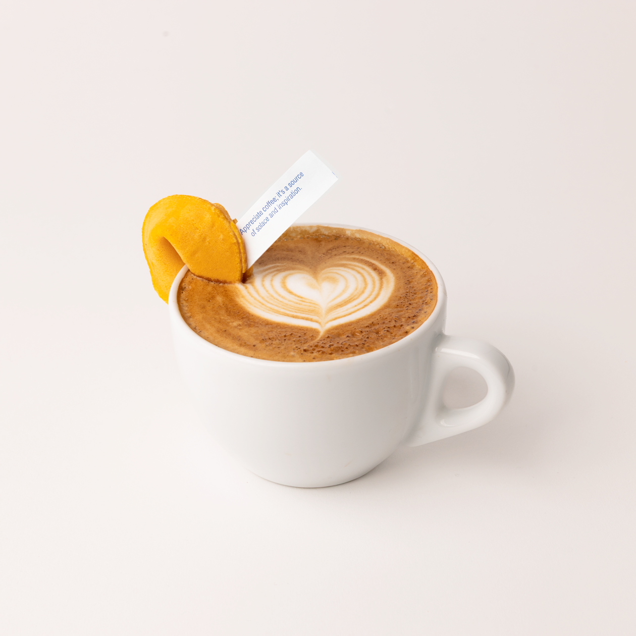 A latte with heart-shaped latte art in a white ceramic mug, garnished with a yellow fortune cookie whose paper fortune reads "Appreciate coffee; it's a source of solace and inspiration."