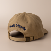 Back of the driftwood brown Frothy Monkey Kong dad hat showing the Frothy Monkey script logo embroidered in navy across the rear panels and the matching cotton adjustable strap with brass clasp