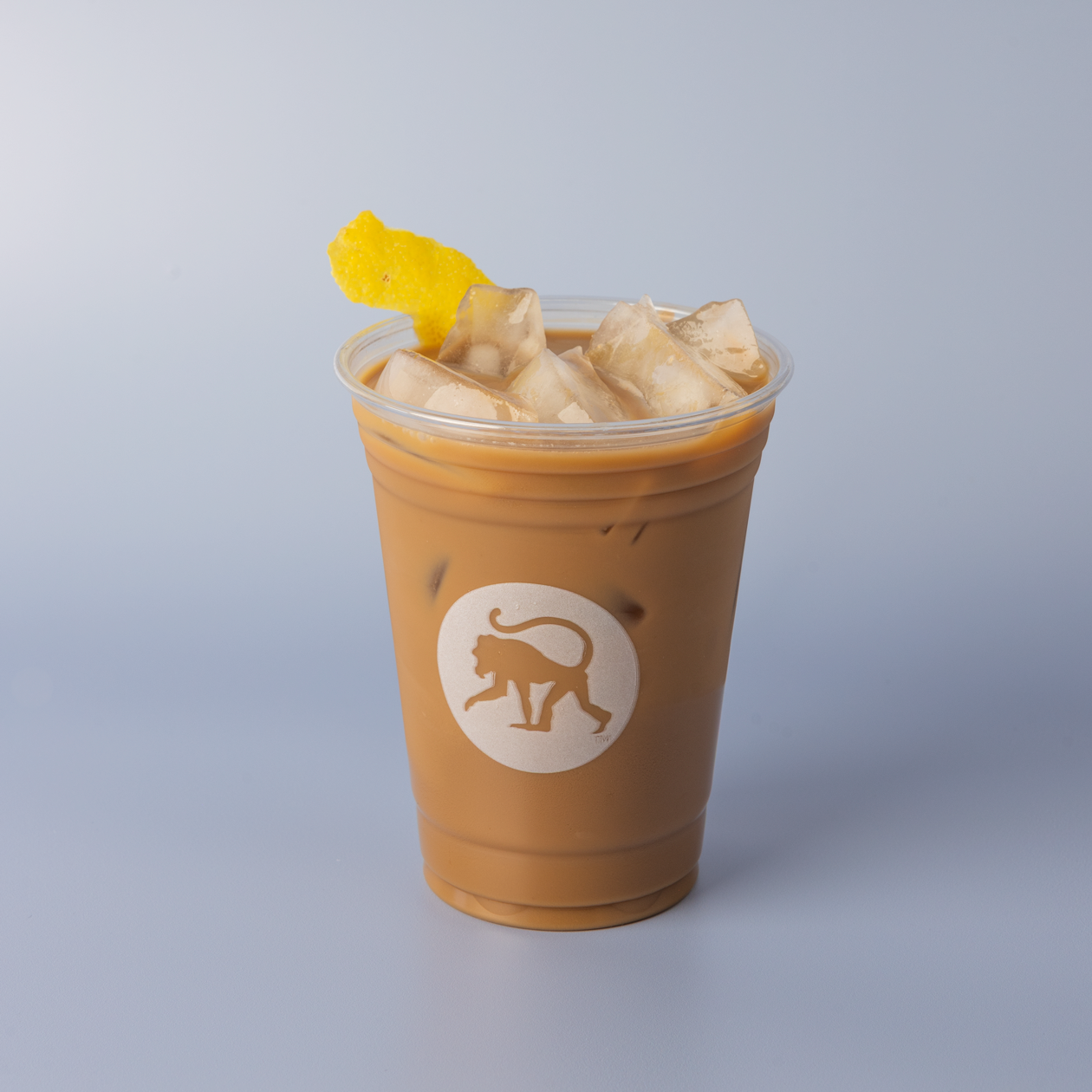 An iced lavender lemon bar cold brew drink in a clear Frothy Monkey cup garnished with a curl of fresh lemon peel, set against a soft blue background.