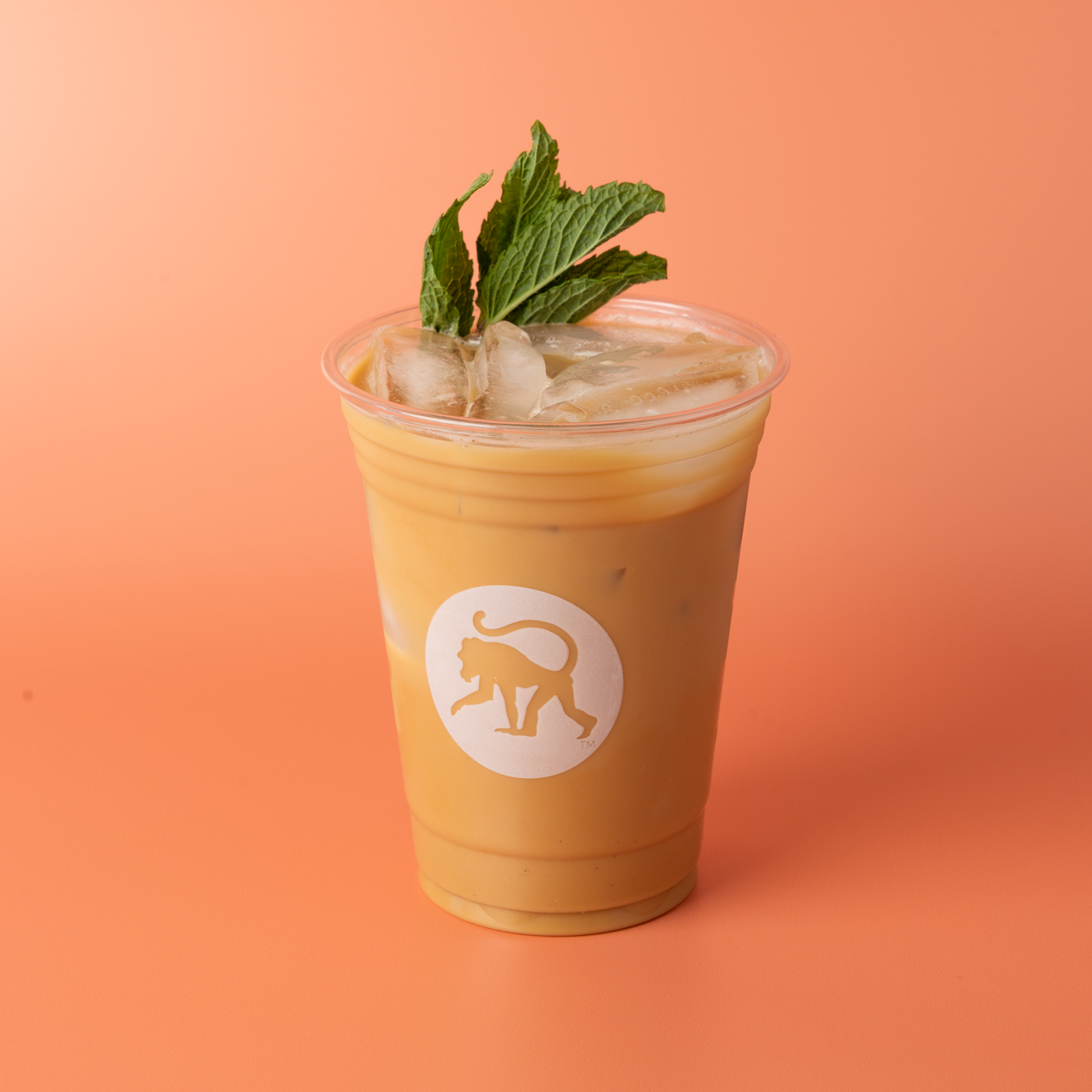 An iced mint julip latte in a clear Frothy Monkey cup garnished with fresh mint sprigs, set against a coral orange background.