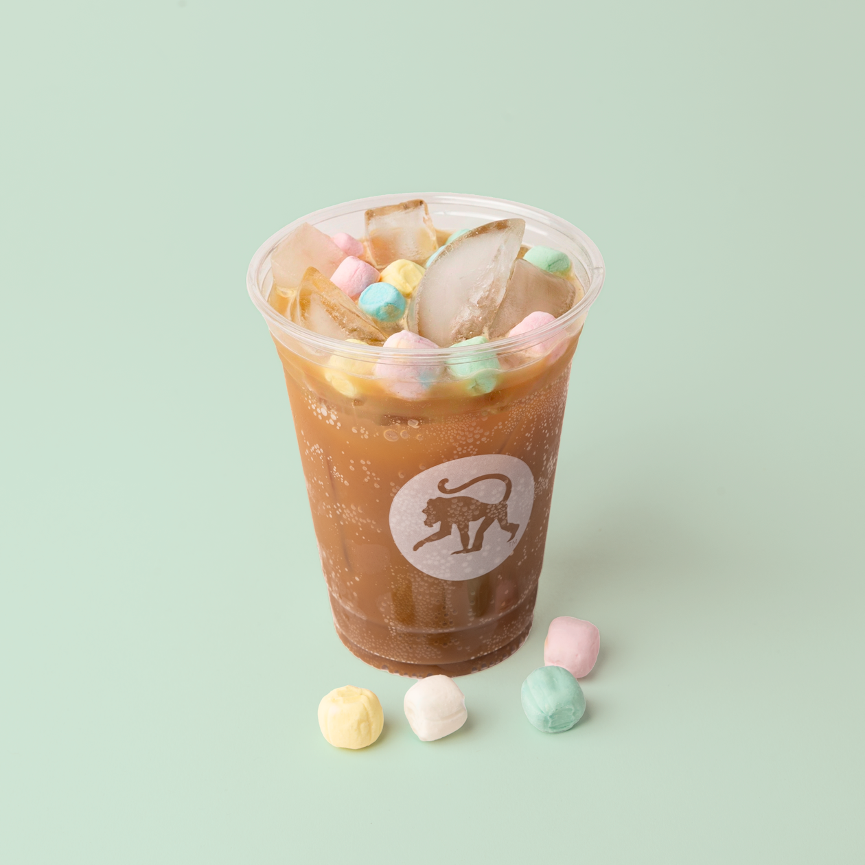 An iced sparkling drink in a clear Frothy Monkey cup topped with pastel buttermint candies, with extra buttermint candies scattered on a mint green surface.