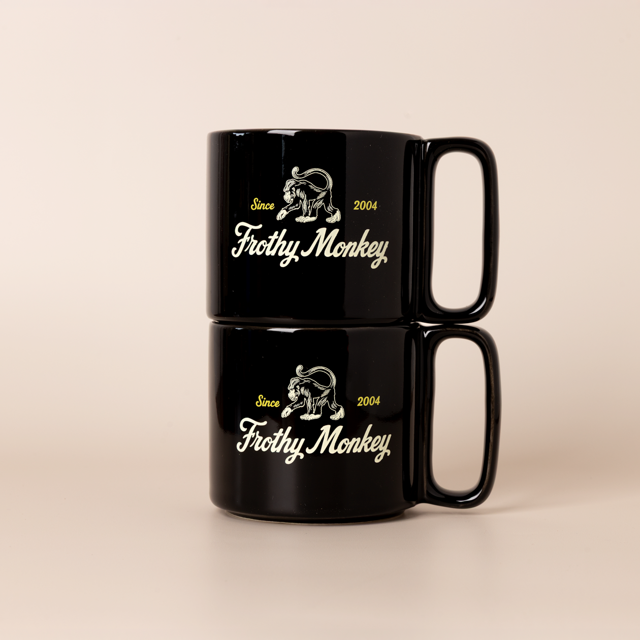 Two Frothy Monkey 10 oz stackable ceramic mugs in Black stacked on top of each other, showing vintage Since 2004 monkey logo in cream with yellow accents