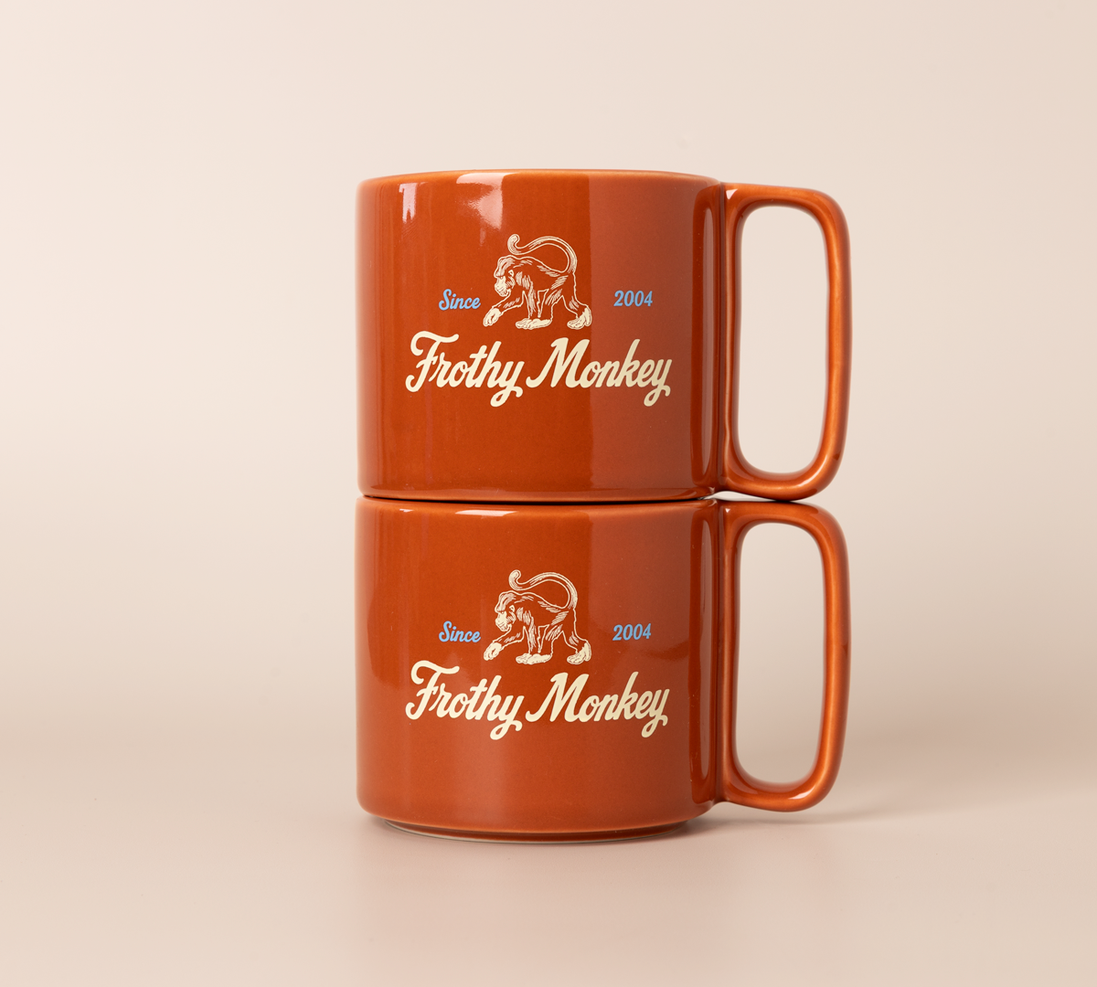 Two Frothy Monkey 10 oz stackable ceramic mugs in burnt orange stacked on top of each other, showing vintage Since 2004 monkey logo in cream with Howler Blue accents