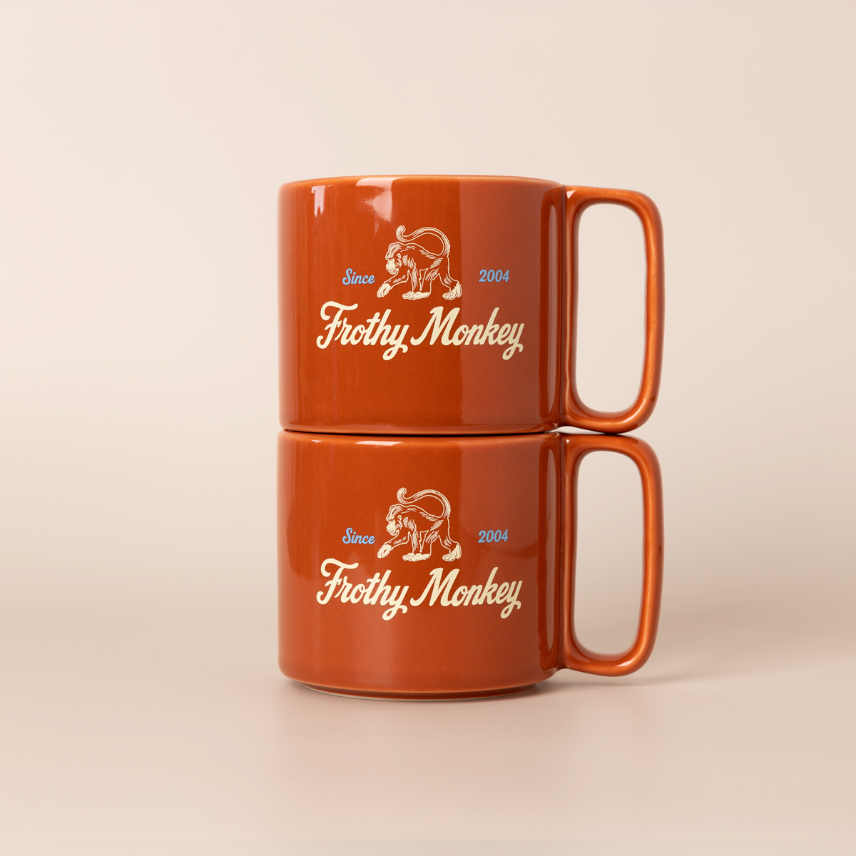 Two Frothy Monkey 10 oz stackable ceramic mugs in burnt orange stacked on top of each other, showing vintage Since 2004 monkey logo in cream with Howler Blue accents