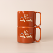 Two Frothy Monkey 10 oz stackable ceramic mugs in burnt orange stacked on top of each other, showing vintage Since 2004 monkey logo in cream with Howler Blue accents