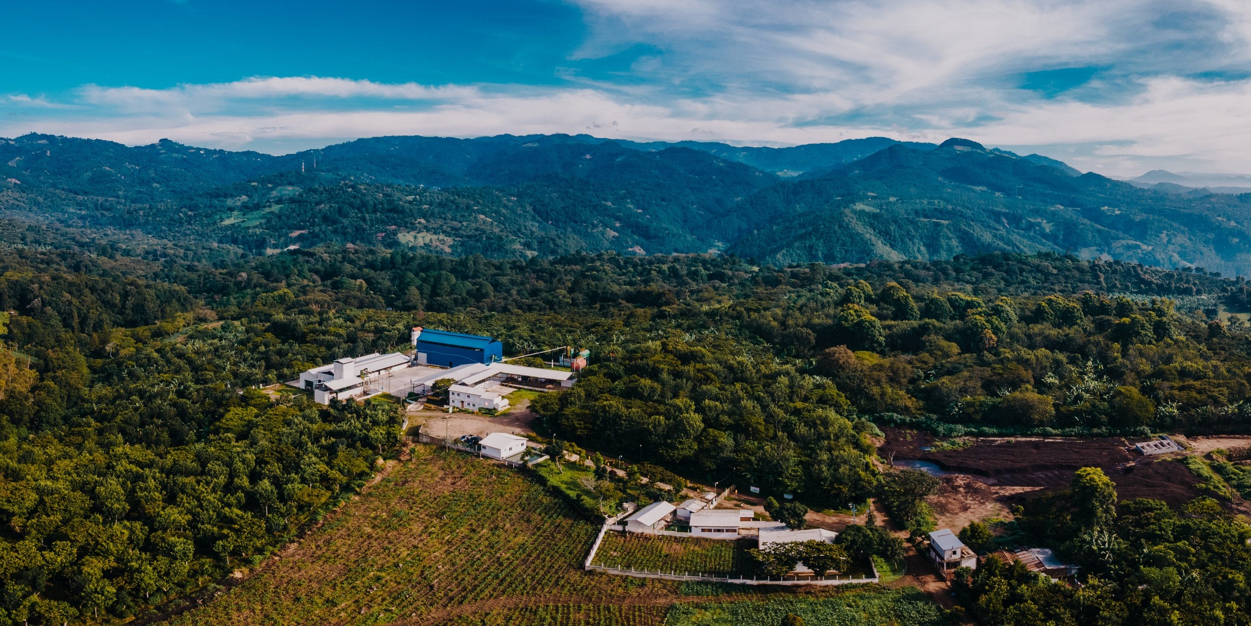 Aerial view of a coffee mill with a blue warehouse surrounded by forested hills, Guatemala