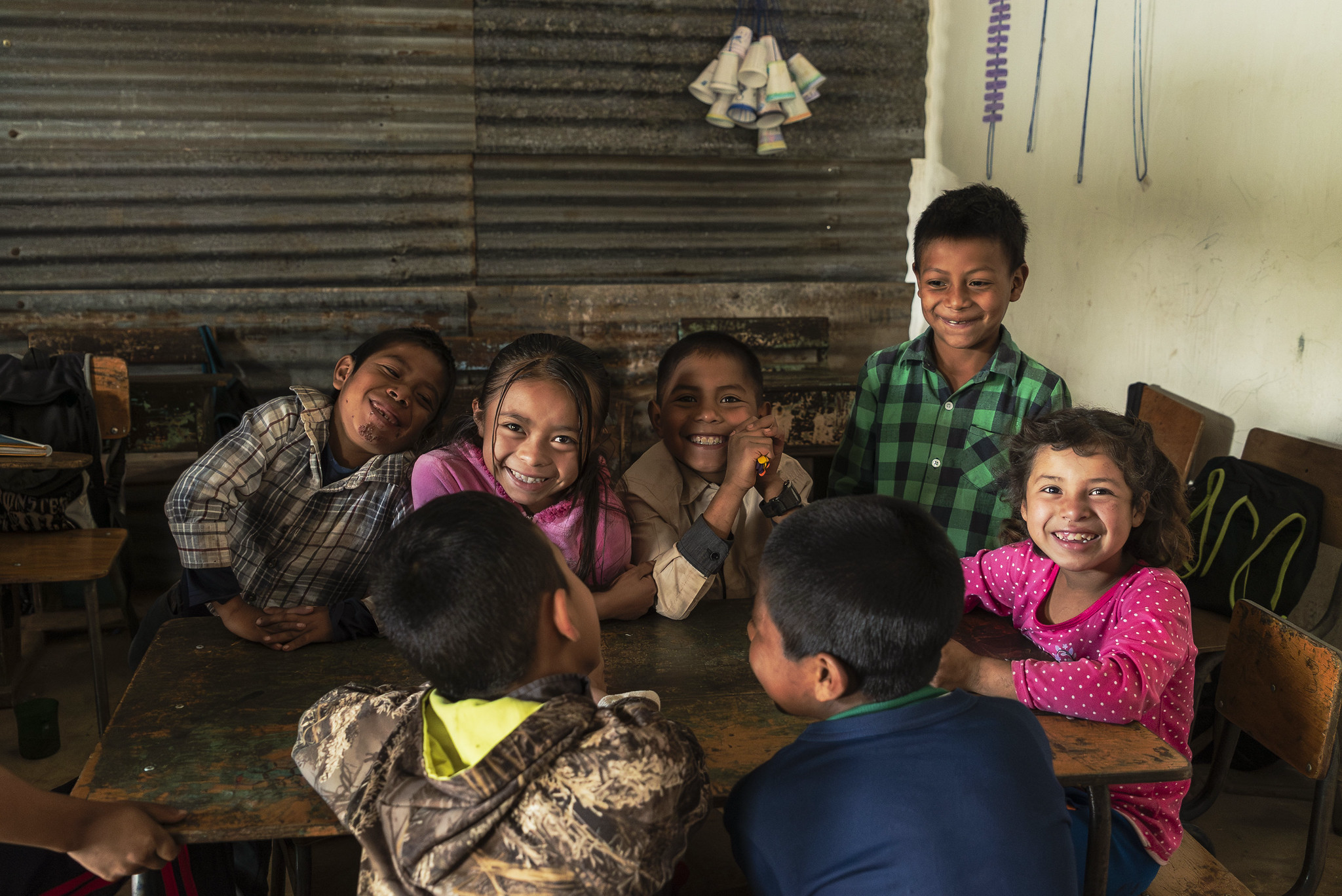 Smiling kids gathered around a wooden desk in a classroom with tin walls, Guatemala