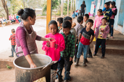 Kids lined up with cups as a woman serves a drink from a large pot outside a school, Guatemala