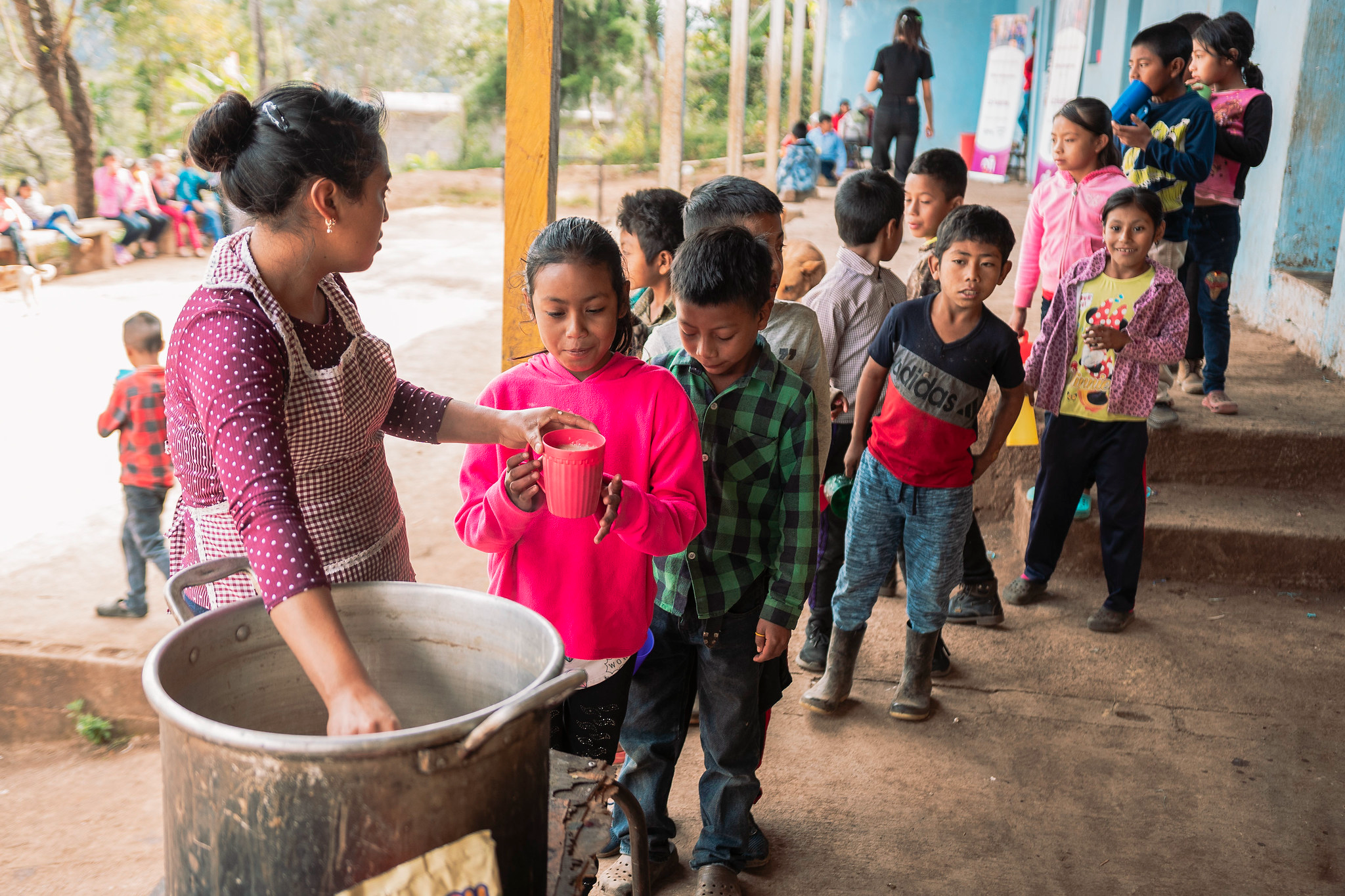 Kids lined up with cups as a woman serves a drink from a large pot outside a school, Guatemala